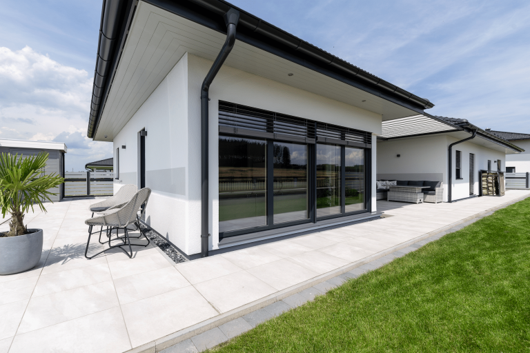 Hartl Haus zeigt einen weißen Bungalow mit schwarzen Fensterrahmen,  großen Glasflächen, einer mit Steinplatten belegten Terrasse und Gartenmöbeln.