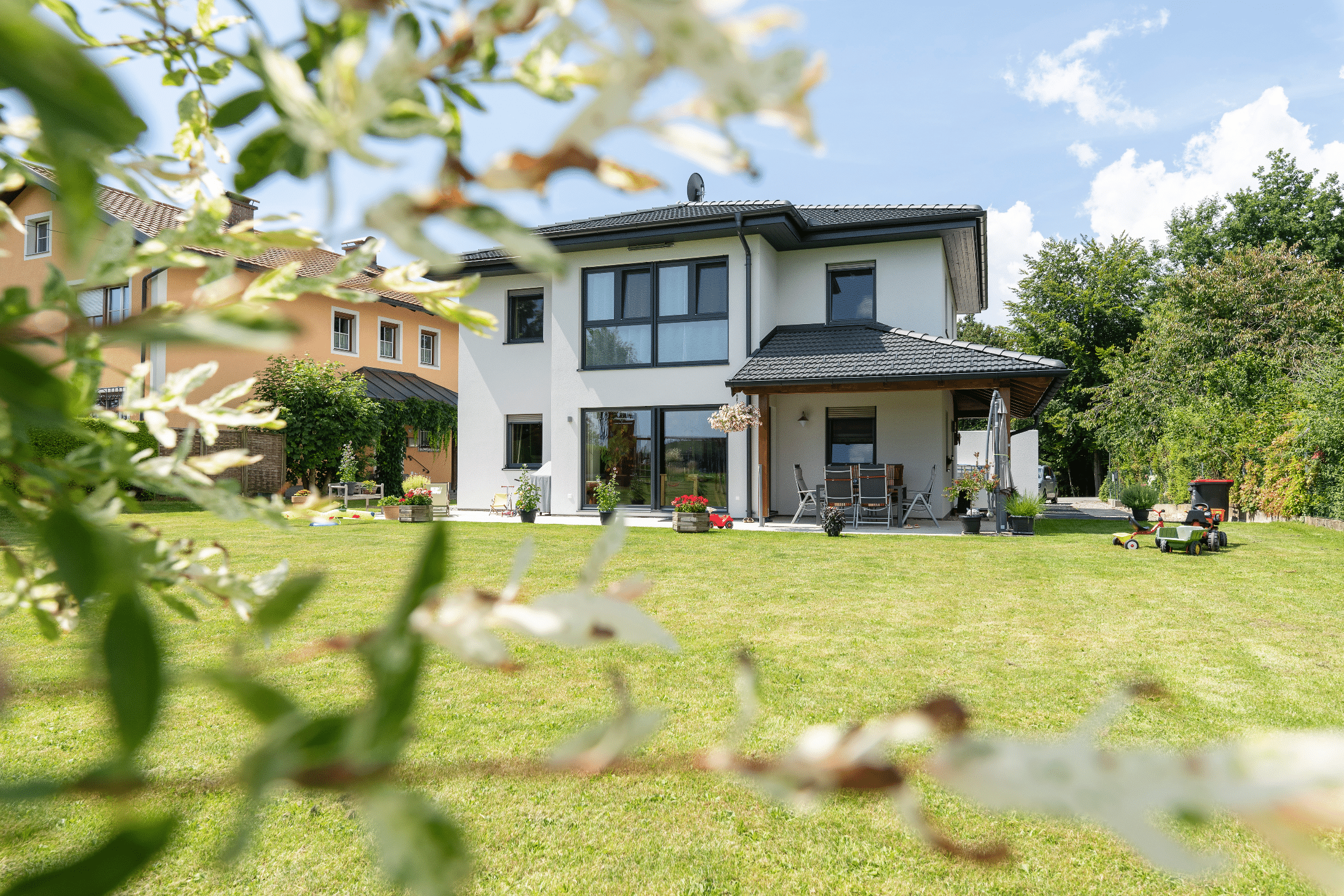 Hartl Haus zeigt ein zweistöckiges Einfamilienhaus in weiß, mit schwarzen Fensterrahmen und Dächern, einer überdachten Terrasse mit Gartenmöbeln und Grünfläche.