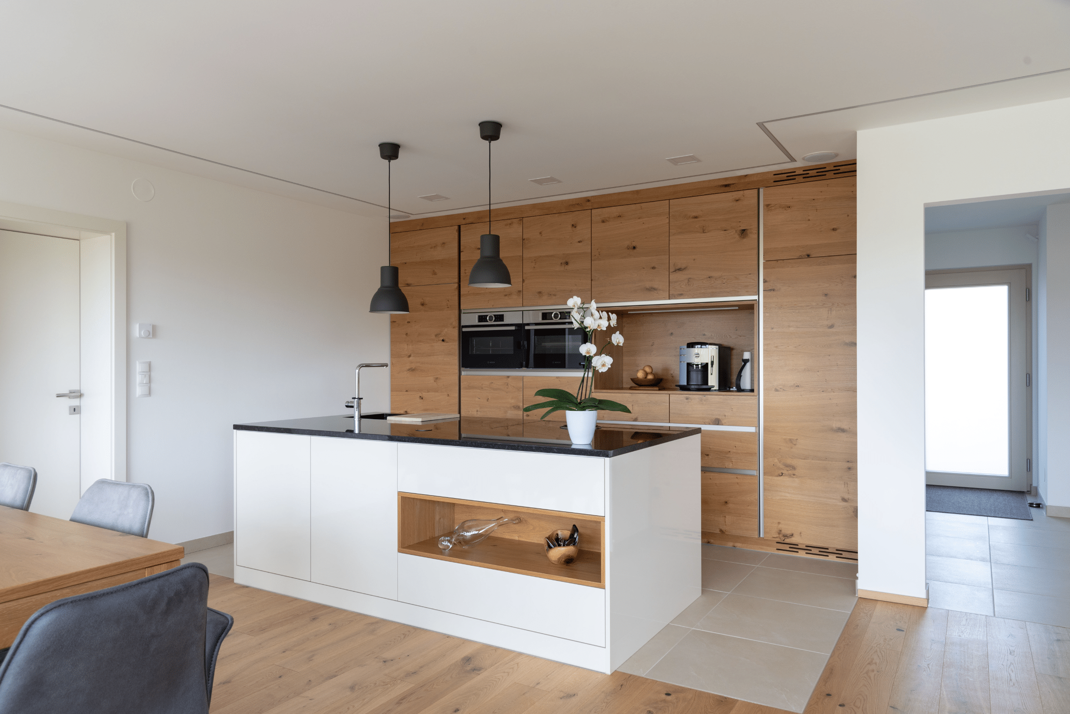 Hartl Haus zeigt eine Küche aus Holz mit weißen Elementen, zwei Backöfen, und schwarzen von der Decke hängenden Lampen.