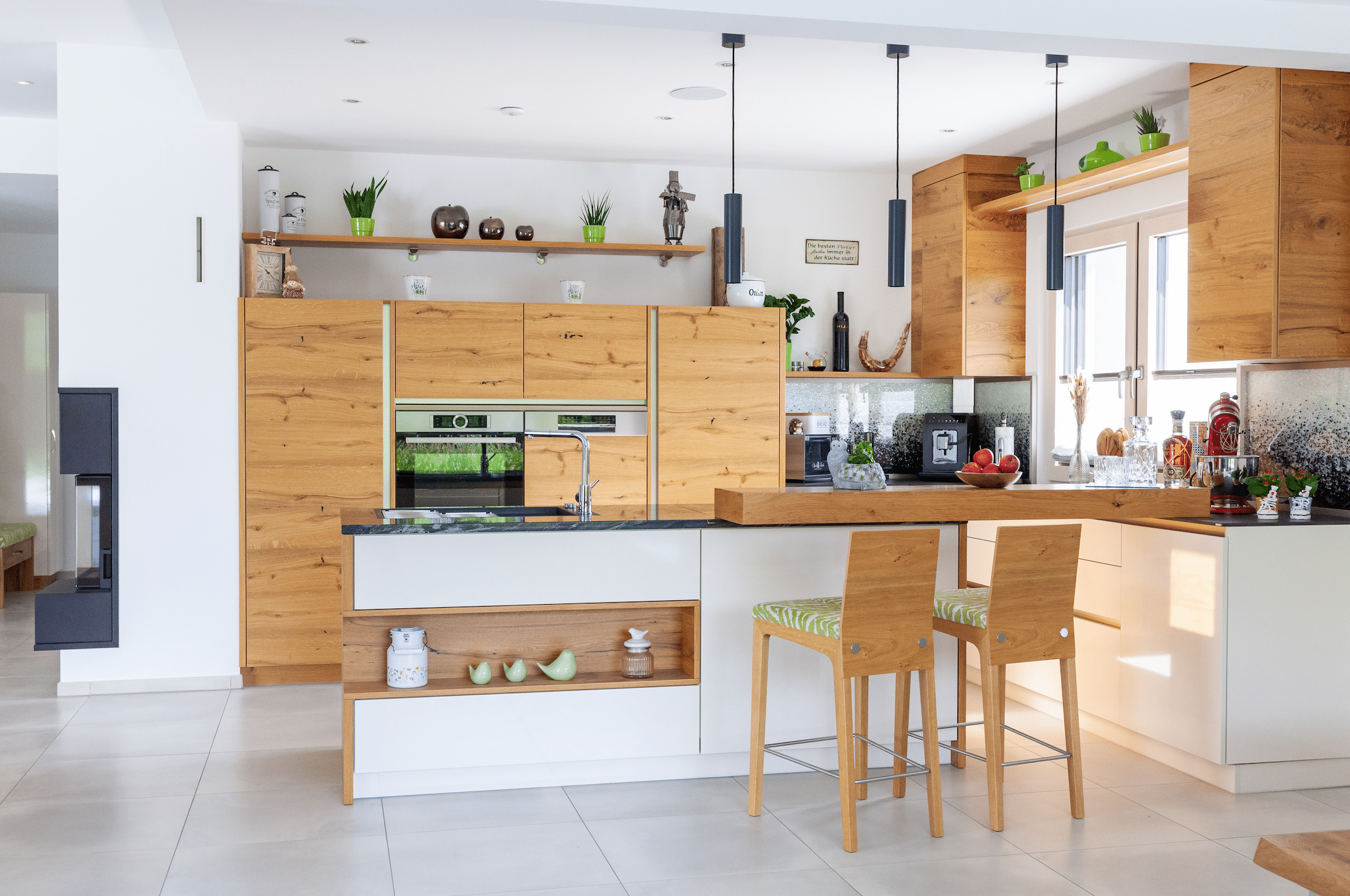 Hartl Haus zeigt eine Küche aus Holz mit verschiedenen Elektrogeräten, einer Barfunktion, hohen Stühlen dazu und weißen Fliesen am Boden.