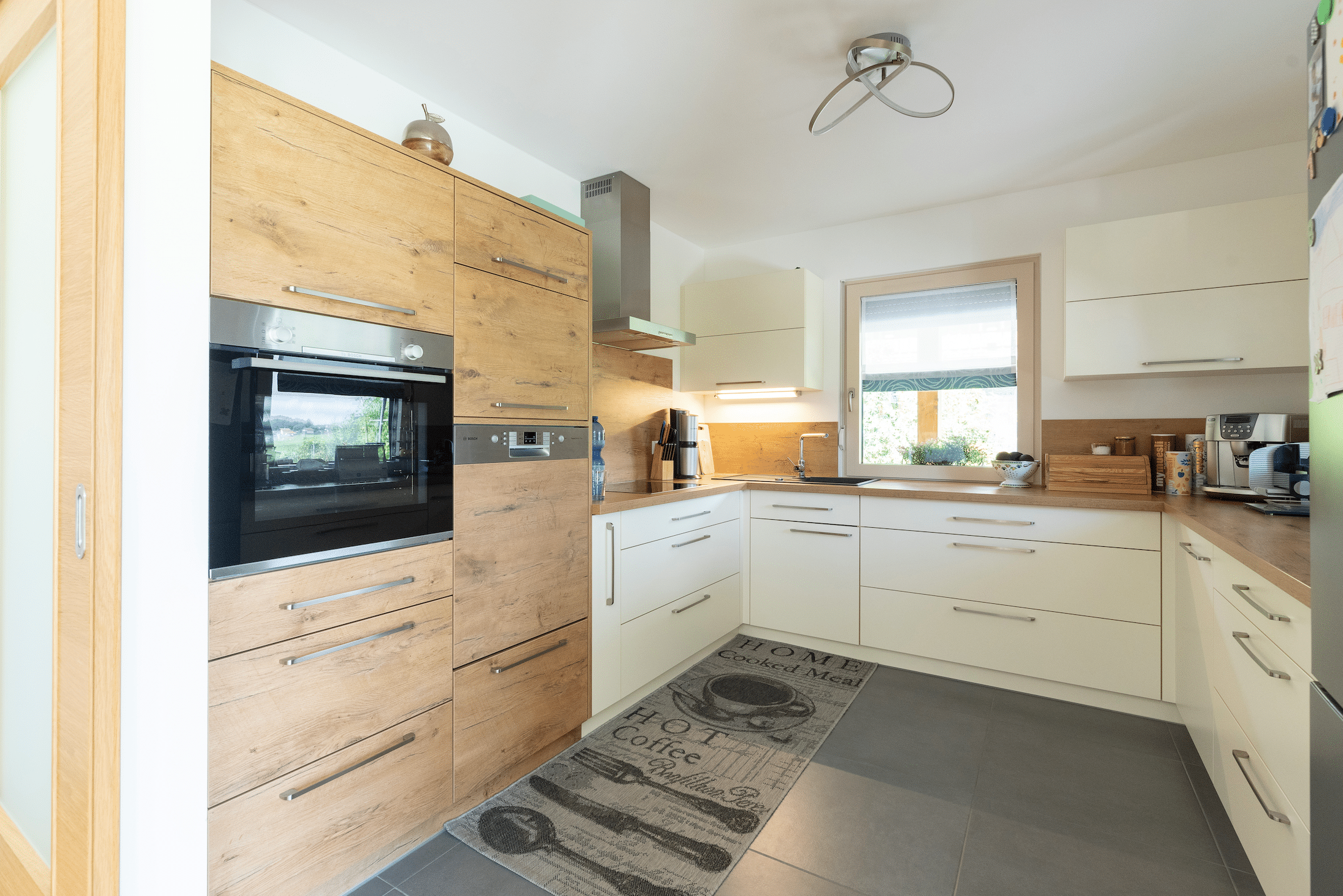 Hartl Haus zeigt eine Küche aus hellem Holz, mit weißen Türen, Teppich und grauen Fliesen am Boden.