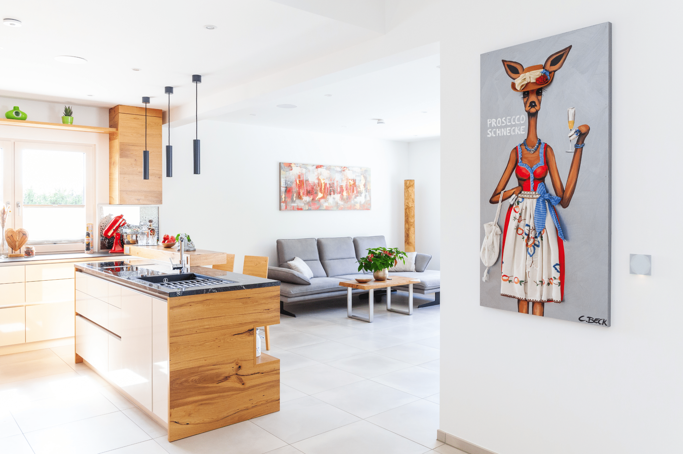 Hartl Haus zeigt eine Küche aus goldigem Holz mit dunkler Arbeitsplatte, direktem Durchgang ins Wohnzimer mit grauer Couch und einem Bild mit modernem Design.