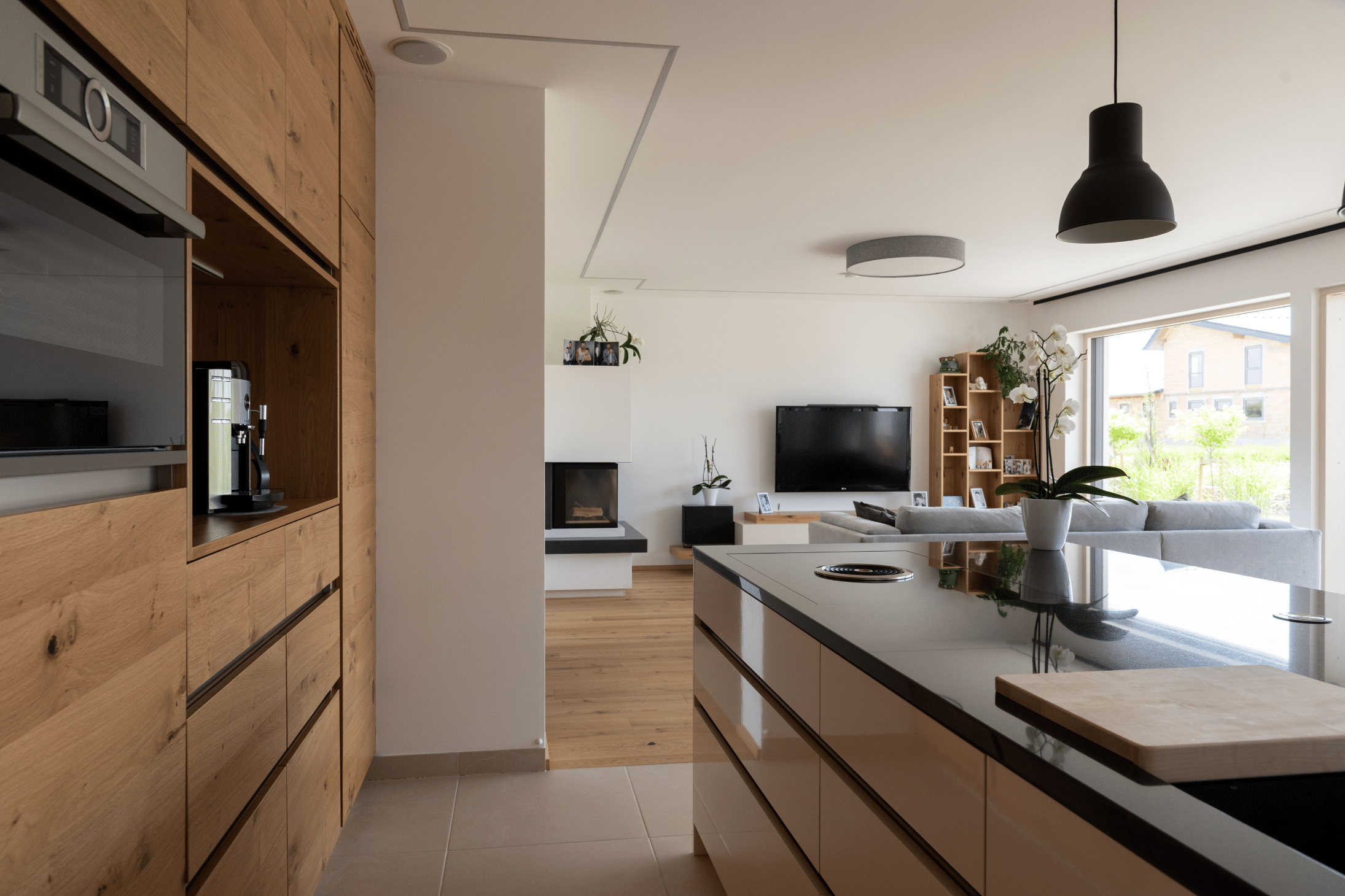 Hartl Haus zeigt eine Küche aus Holz mit weißen Schranktüren, mehreren Elektrogeräten, einer schwarzen von der Decke hängenden Lampe und Terrassentür.