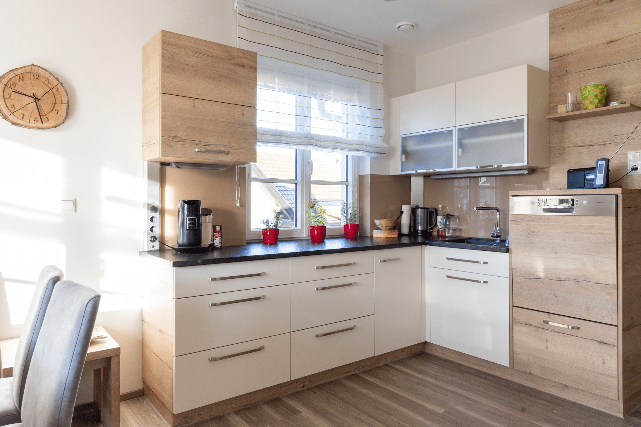 Hartl Haus zeigt eine Küche aus Holz mit weißen Elementen, einem Fenster mit Vorhängen und Esstisch.