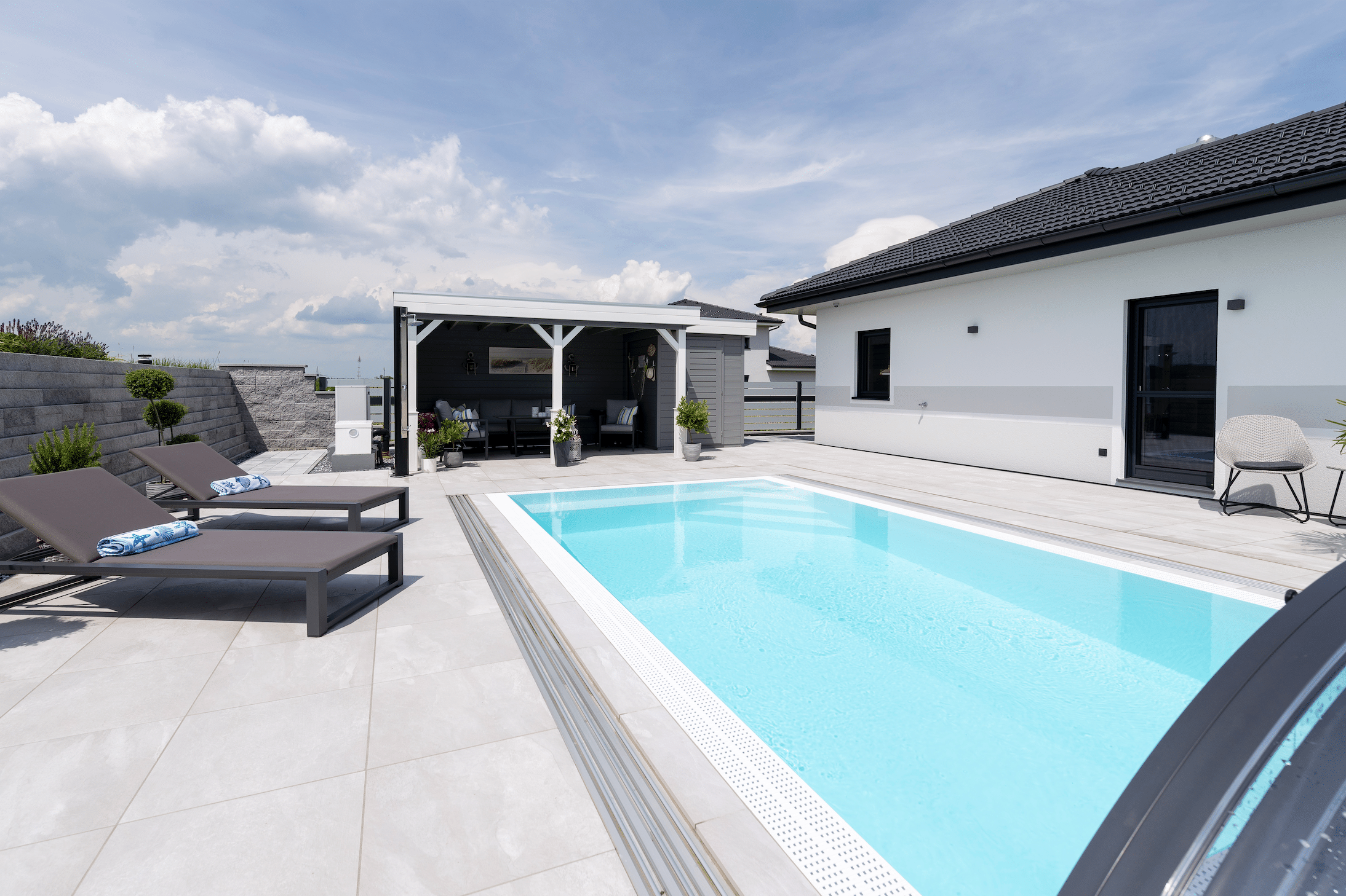 Hartl Haus zeigt eine Terrasse mit großen hellgrauen Fliesen, einen rechteckigen Pool mit türkisem Wasser, Liegestühle und einer überdachten Lounge.