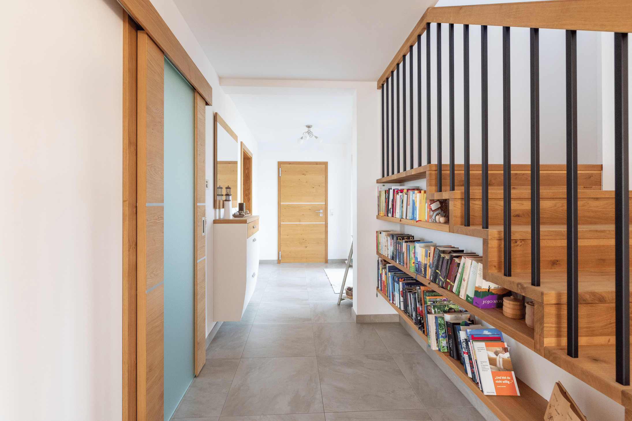 Hartl Haus zeigt ein Vorhaus mit grauen Fliesen am Boden, Holzteppen und einem Bücherregal darunter.