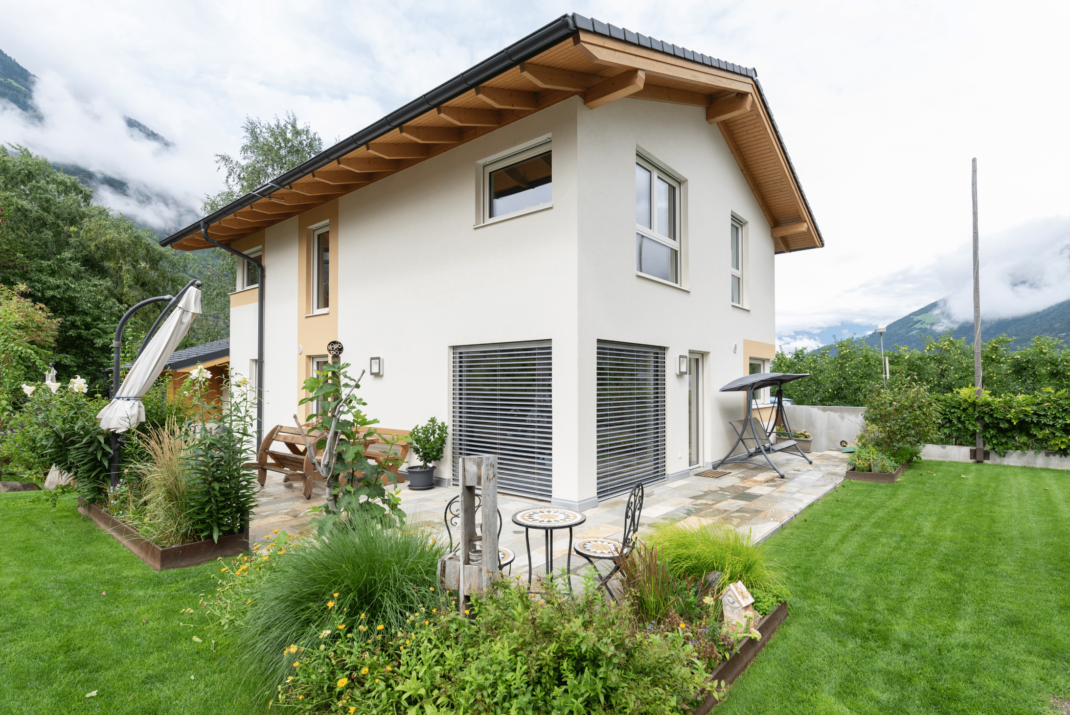 Hartl Haus zeigt ein weißes Einfamilienhaus mit Satteldach, einer Terrasse mit verschiedenen Gartenmöbeln und einem bepflanzten Garten.