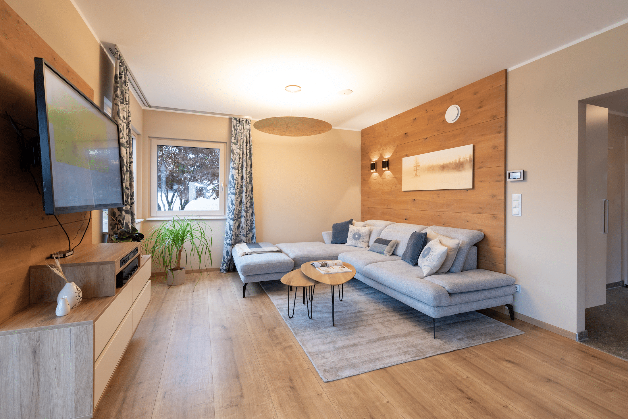 Hartl Haus zeigt ein Wohnzimmer mit Holz verbaut, hellgrauer Couch, einem Couchtisch, gemusterten Vorhängen und einem an der Wand hängenden Fernseher.