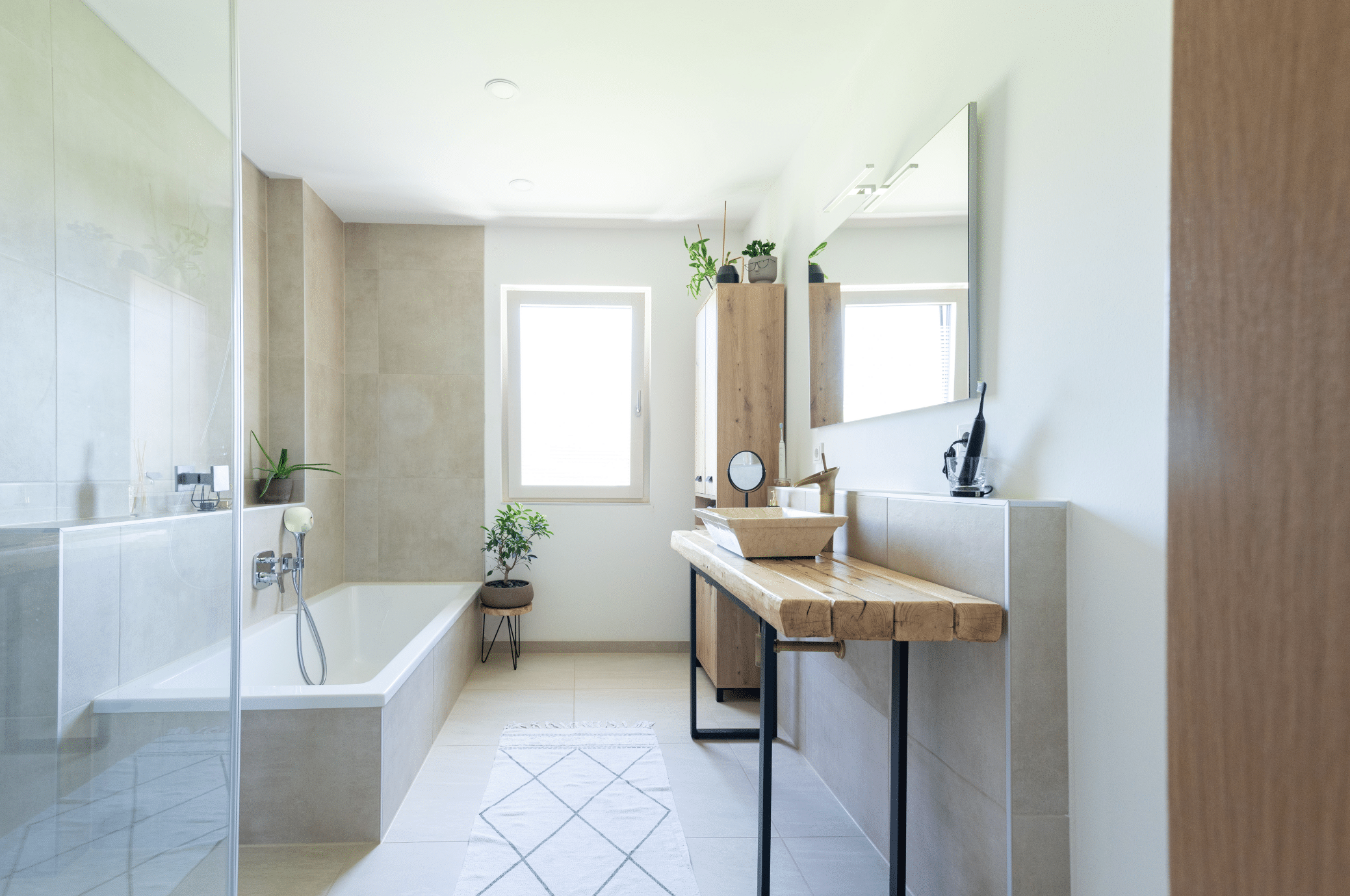 Hartl Haus zeigt ein hell gefliestes Badezimmer mit eckiger Badewanne, silbernen Armaturen und einen Waschtisch aus Holz und Hochschrank.