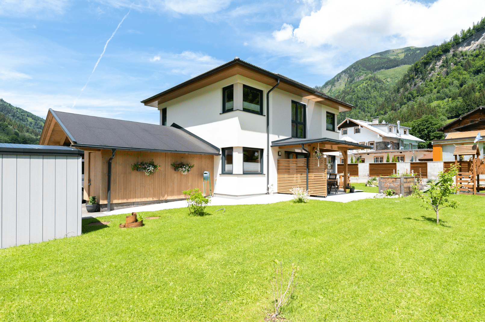Hartl Haus zeigt ein helles Einfamilienhaus im Grünen, mit einem Spielplatz aus Holz, einem Hochbeet, überdachter Terrasse und einem Carport.