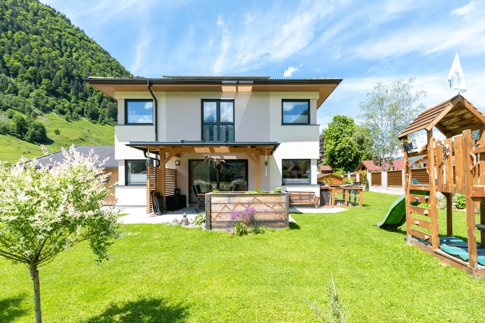 Hartl Haus zeigt ein helles Einfamilienhaus im Grünen, mit einem Spielplatz aus Holz und grüner Rutsche, einem Hochbeet und überdachter Terrasse.