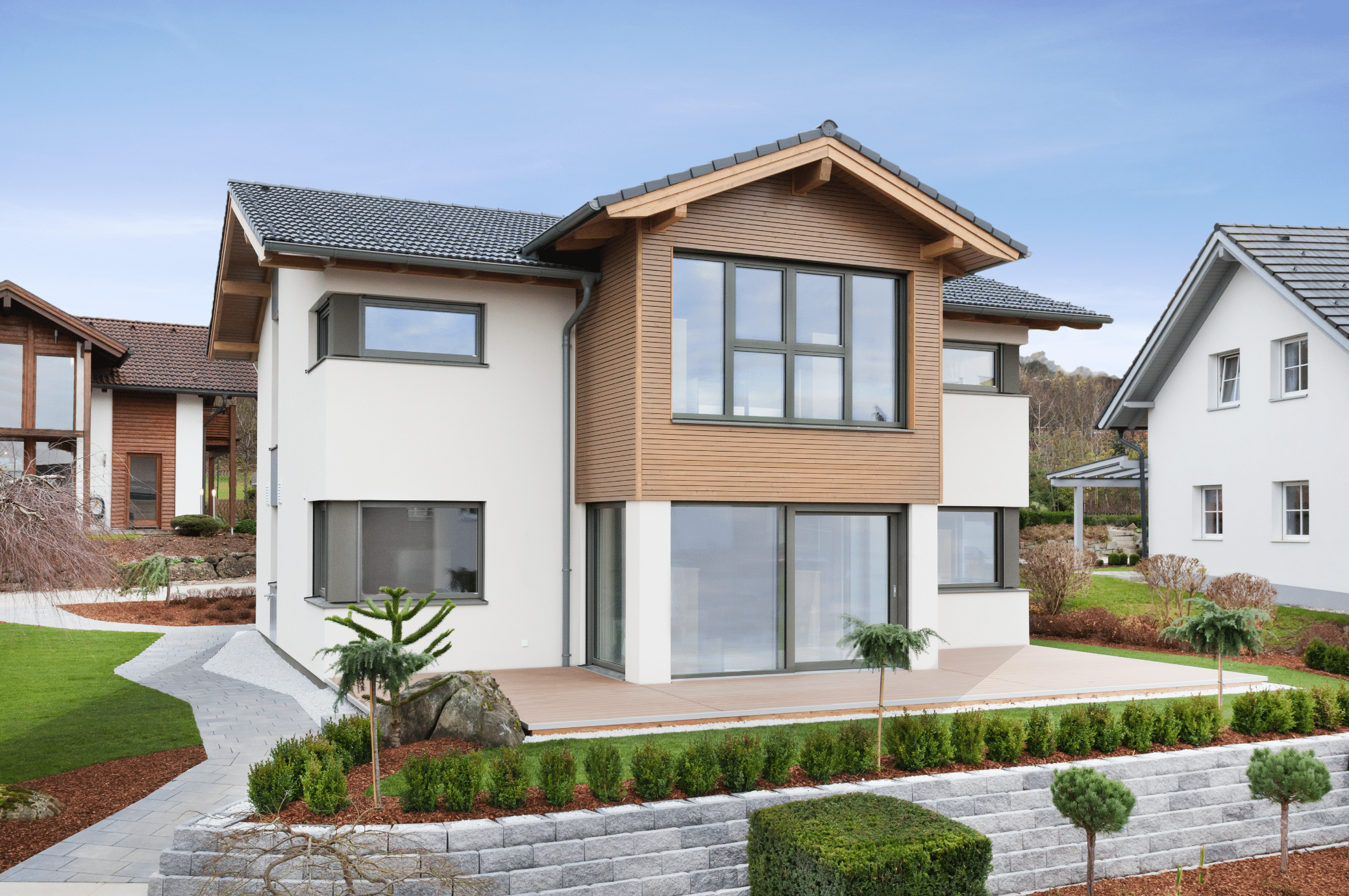Hartl Haus zeigt ein helles Einfamilienhaus mit Holzverkleidung, Terrasse und gepflegtem Vorgarten mit Zugang zum Haus aus Steinplatten und niedriger Mauer.