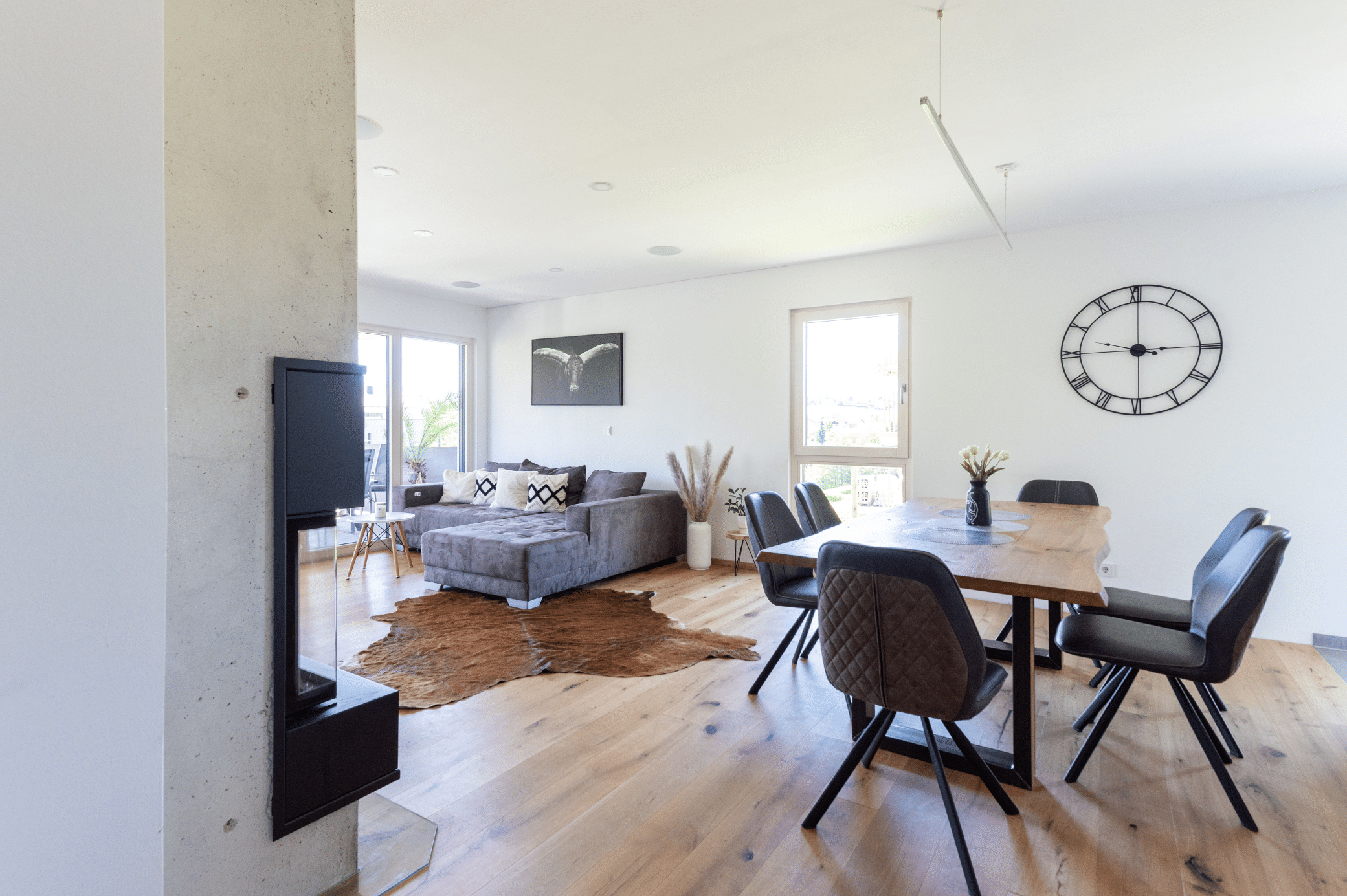 Hartl Haus zeigt eine offene Wohnküche mit Kamin, einem Esstisch aus Holz, Stühlen und Blick auf die graue Couch mit braunem Tierfell am Boden.