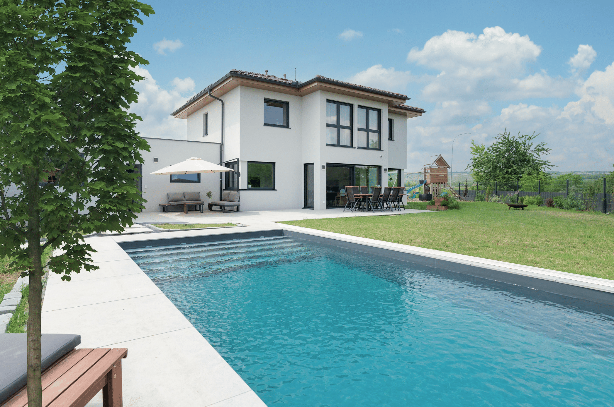 Hartl Haus zeigt ein weisses Einfamilienhaus mit Wintergarten und Terrasse, einer Gartenlounge mit grauen Sitzbänken und einem rechteckigen Pool.