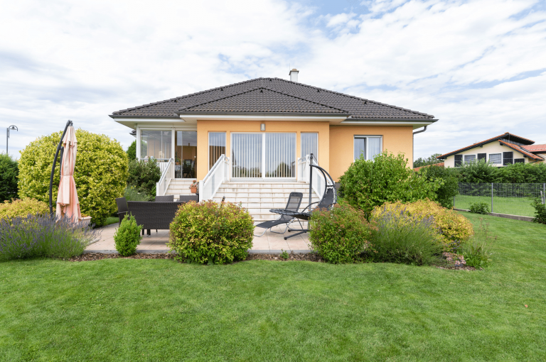 Hartl Haus zeigt ein Einfamilienhaus in orange mit einem schwarzen Dach, großen Fenstern und einer Terrassentür mit Zugang über Stufen zur großen Terrasse mit Loungemöbeln und einem Sonnenschirm.