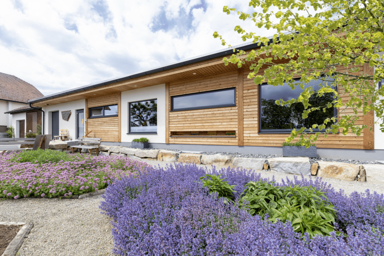 HARTL HAUS zeigt den Vorgarten eines Bungalows in weiß mit Holzverbau, ein quadratisches Fenster, eine Steinmauer und Bänkchen.