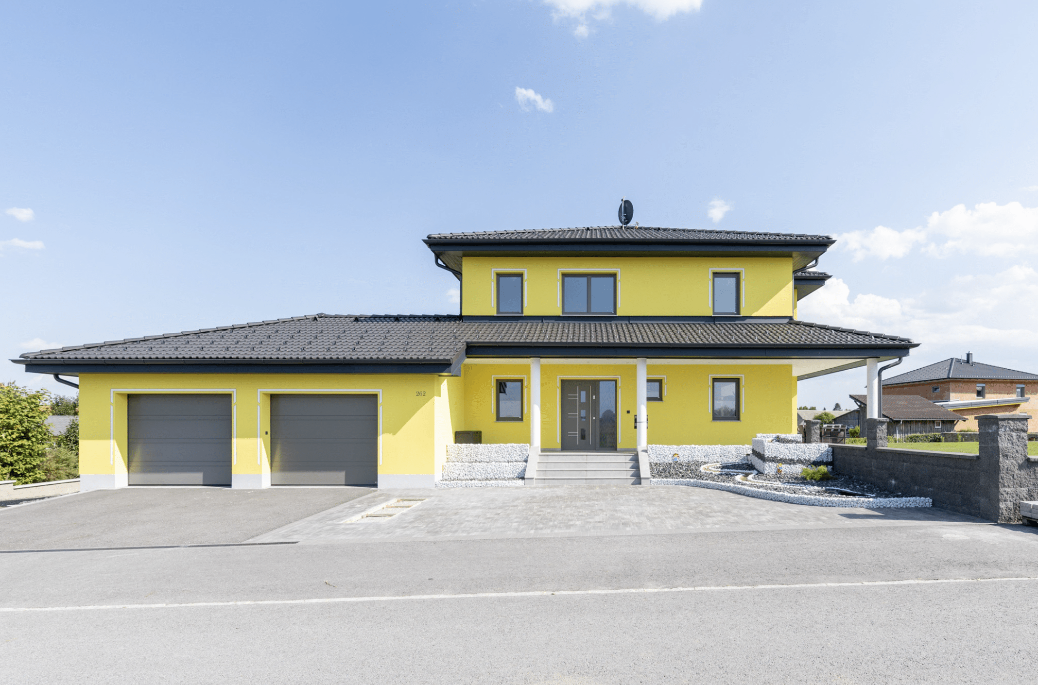 HARTL HAUS zeigt ein gelbes, zweistöckiges Einfamilienhaus mit Doppelgarage und geräumiger Zufahrt.