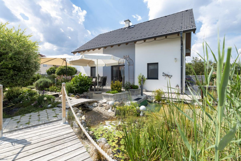 Einfamilienhaus mit Satteldach, grauen Farbakzenten, dunkelgraue Alufensterrahmen, romantisch angelegter Terrasse und großem Biotop von Hartl Haus.