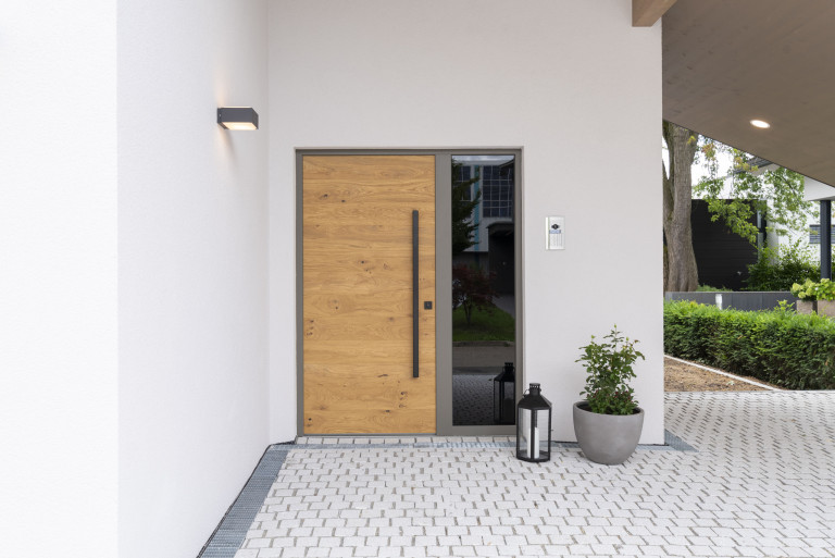 Hartl Haus zeigt weißes Haus mit Holzeingangstüre mit Glas-Seitenteil und grauem Türrahmen.