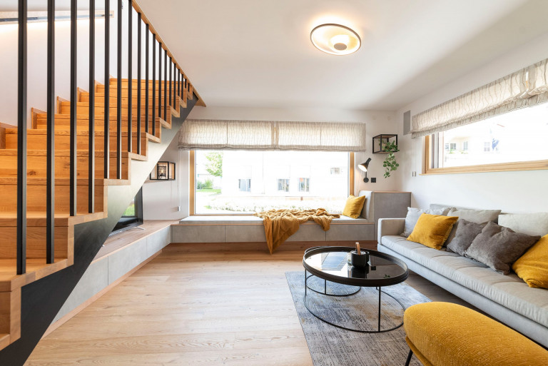 Hartl Haus zeigt gemütliches Wohnzimmer mit langer grauer Couch und großer gepolsterter Fensterbank vor großer Fensterfläche mit Blick auf den Stiegenaufgang.