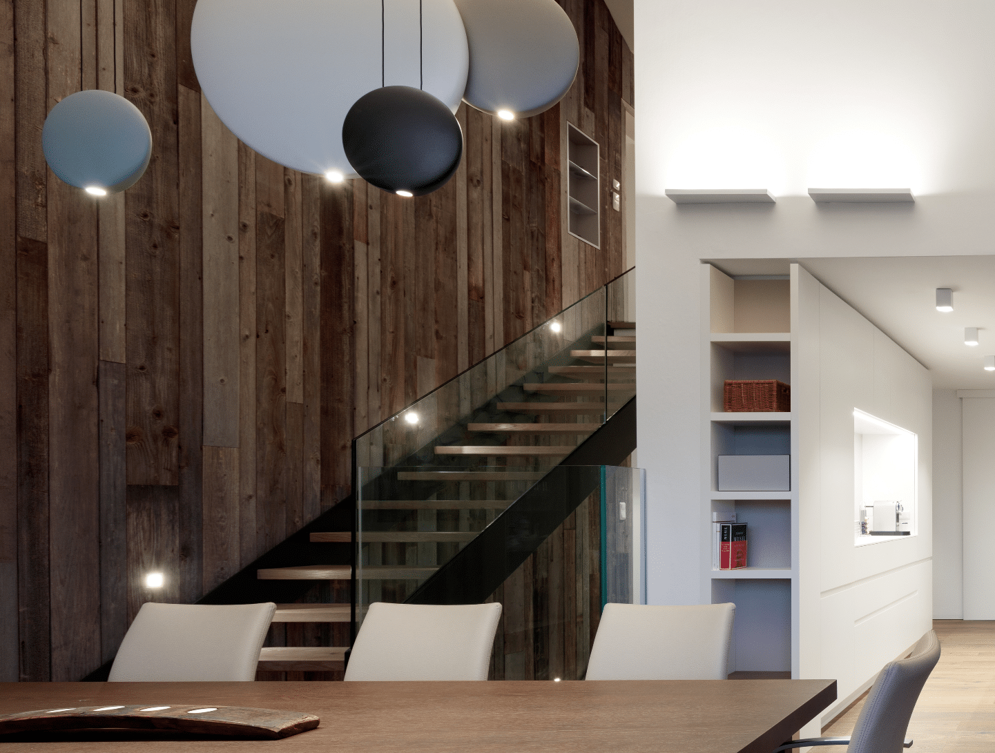 Hausjell zeigt einen Wohnbereich mit Holztisch und hellen Stühlen mit einer dunklen Treppe mit Beleuchtung und Glasgeländer.