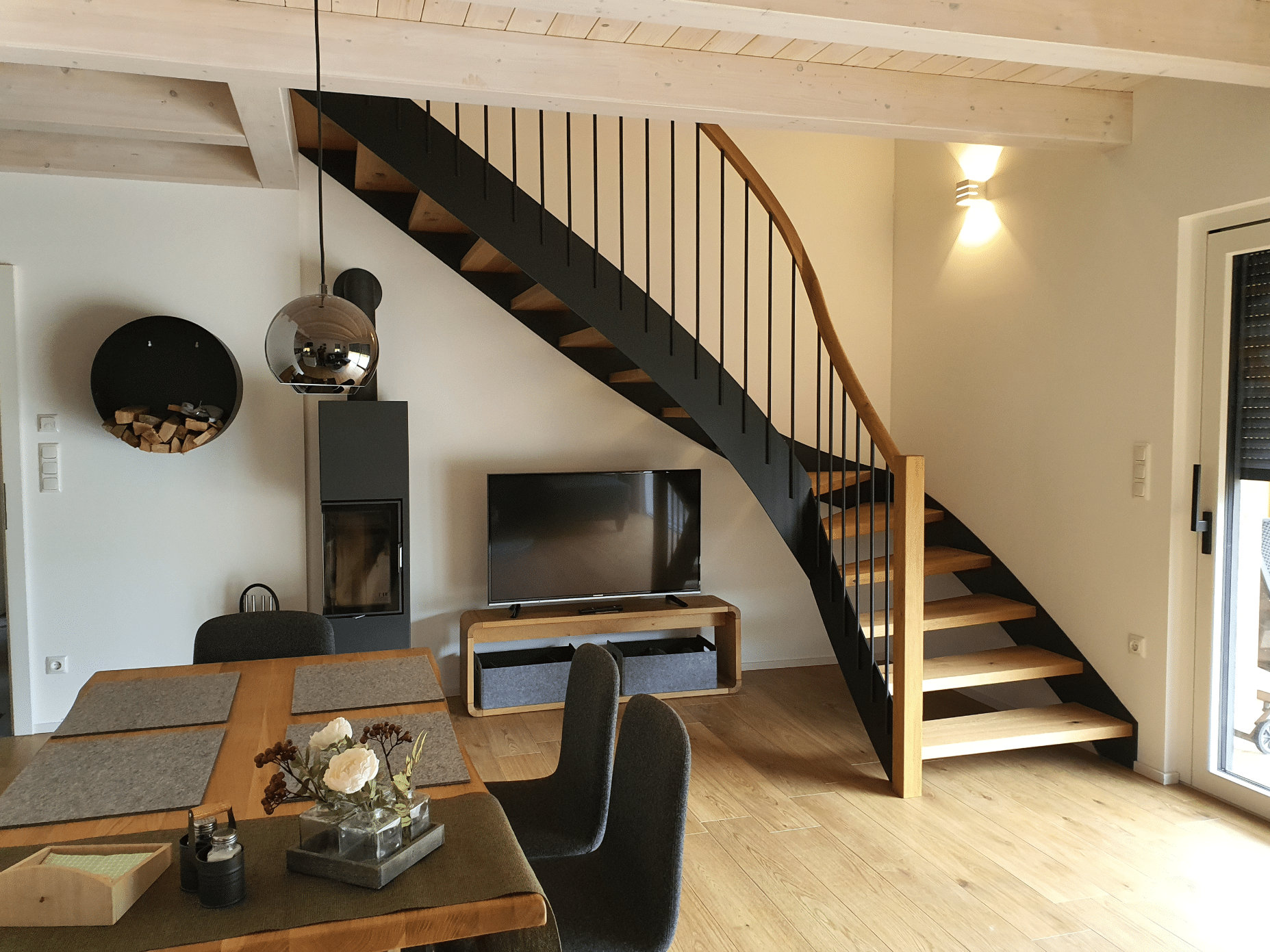 Hausjell zeigt ein Esszimmer mit Holztisch und schwarzen Stühlen, einem Kamin und einer Holztreppe mit schwarzem Geländer und Beleuchtung.