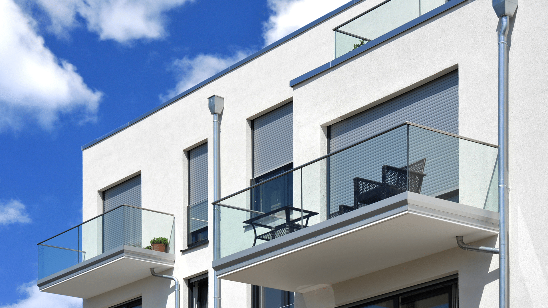 Hella präsentiert dunkelgraue Rollläden als Sonnenschutz am Balkon eines weißen Hauses mit schwarzer Sitzgarnitur.