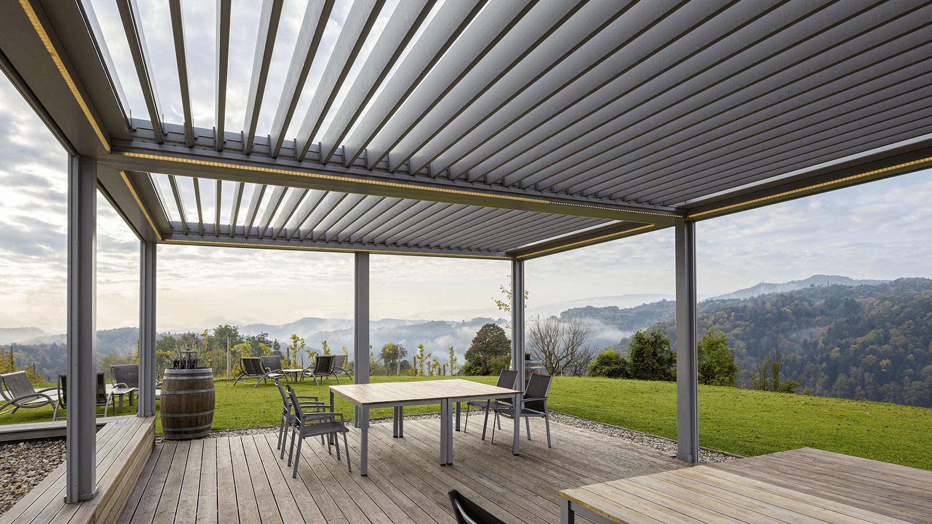 Hella präsentiert eine graue, beleuchtete Pergola-Terrassenüberdachung, darunter befinden sich Holztische und Stühle auf einem Holzboden, mit weiter Aussicht in di Berge.