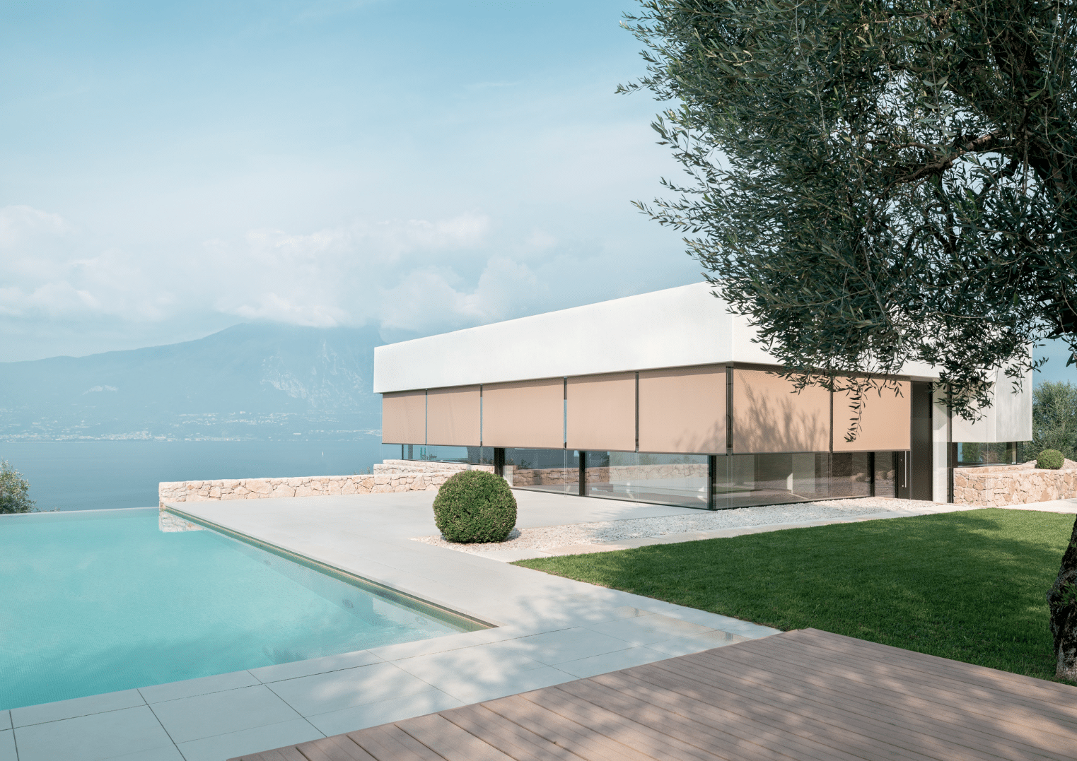 Hella präsentiert beige Senkrechtmarkisen eines Poolhauses mit großen Fensterflächen mit Terrasse davor und einer Grünfläche, gefliester Weg zum Pool, Aussicht auf See und in die Berge.