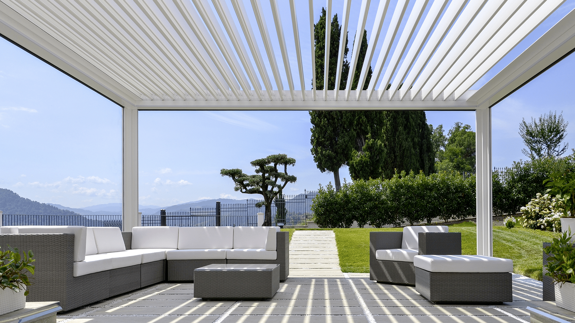 Hella präsentiert eine weiße Pergola-Terrassenüberdachun, darunter Rattanmöbel mit weißen Auflagepolstern in einem schön bepflanztem Garten und strahlend blauem Himmel.
