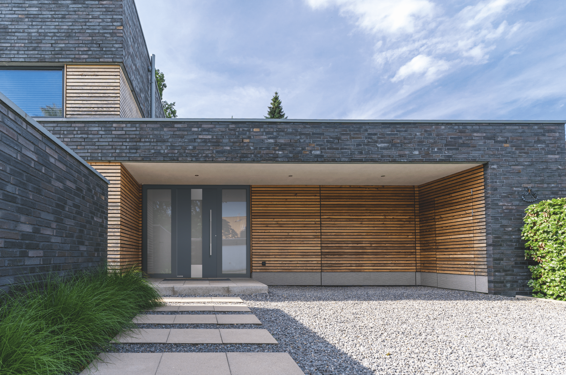 HÖRMANN zeigt ein modernes Haus mit Holzverkleidung, Begrünung im Vorgarten und Steinplatten als Zugang zur grauen Haustüre mit silbernem Griff und Sichtfenstern.