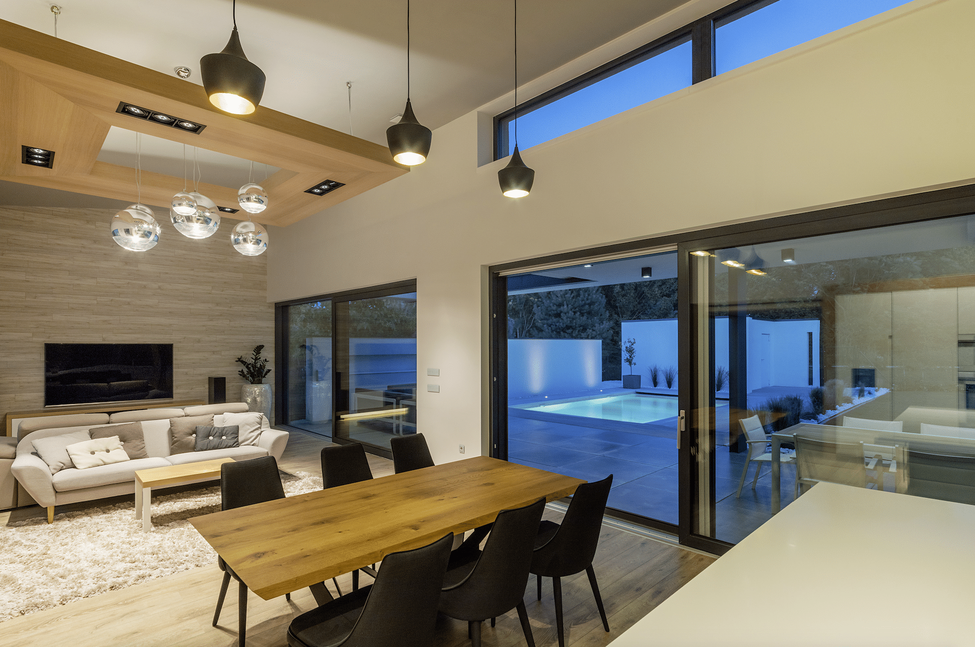 Offenes Wohnkonzept mit gemütlichem Wohnzimmer und Essbereich. Mit Blick durch das Ganzglassystem von Internorm auf die Terrasse und den beleuchteten Pool.