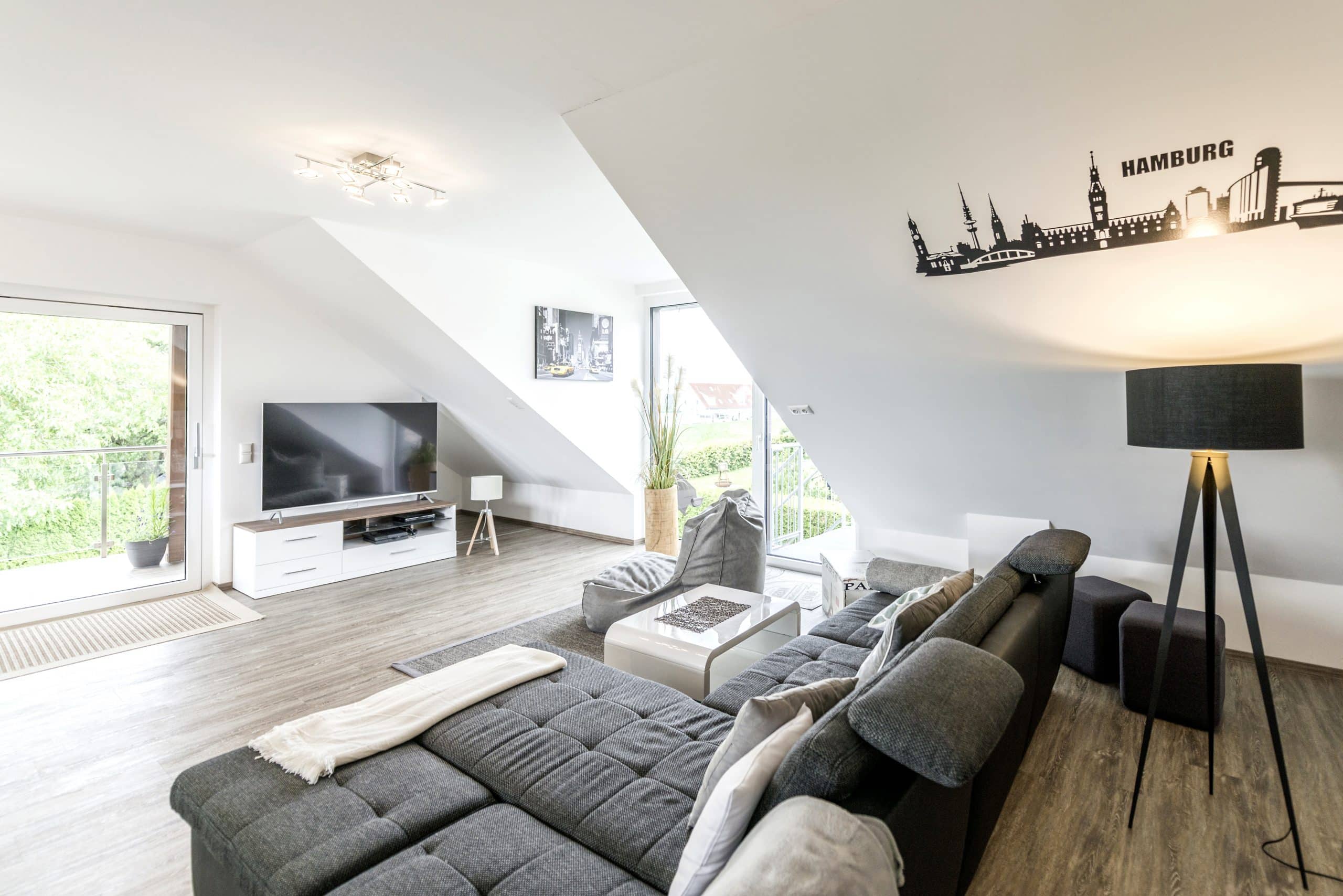 Internorm zeigt einen Wohnraum mit grauer, großflächiger Couch, einem Fernseher, einer Stehlampe und großen Balkontüren.