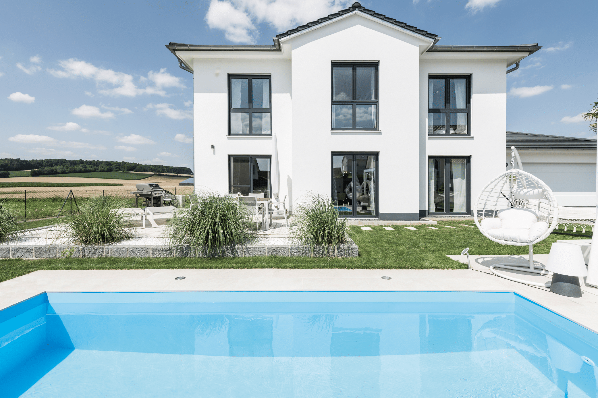 Internorm zeigt ein weisses Einfamilienhaus, große Fenster mit schwarzen Rahmen und eine begrünte Terrasse und eckigem Pool mit weissem Hängesessel.