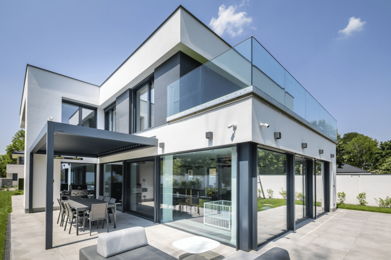 Internorm zeigt ein weisses Einfamilienhaus mit Balkon, Glasgeländer und eine Terrasse mit Überdachung in grau, mit langem Esstisch und Stühlen.