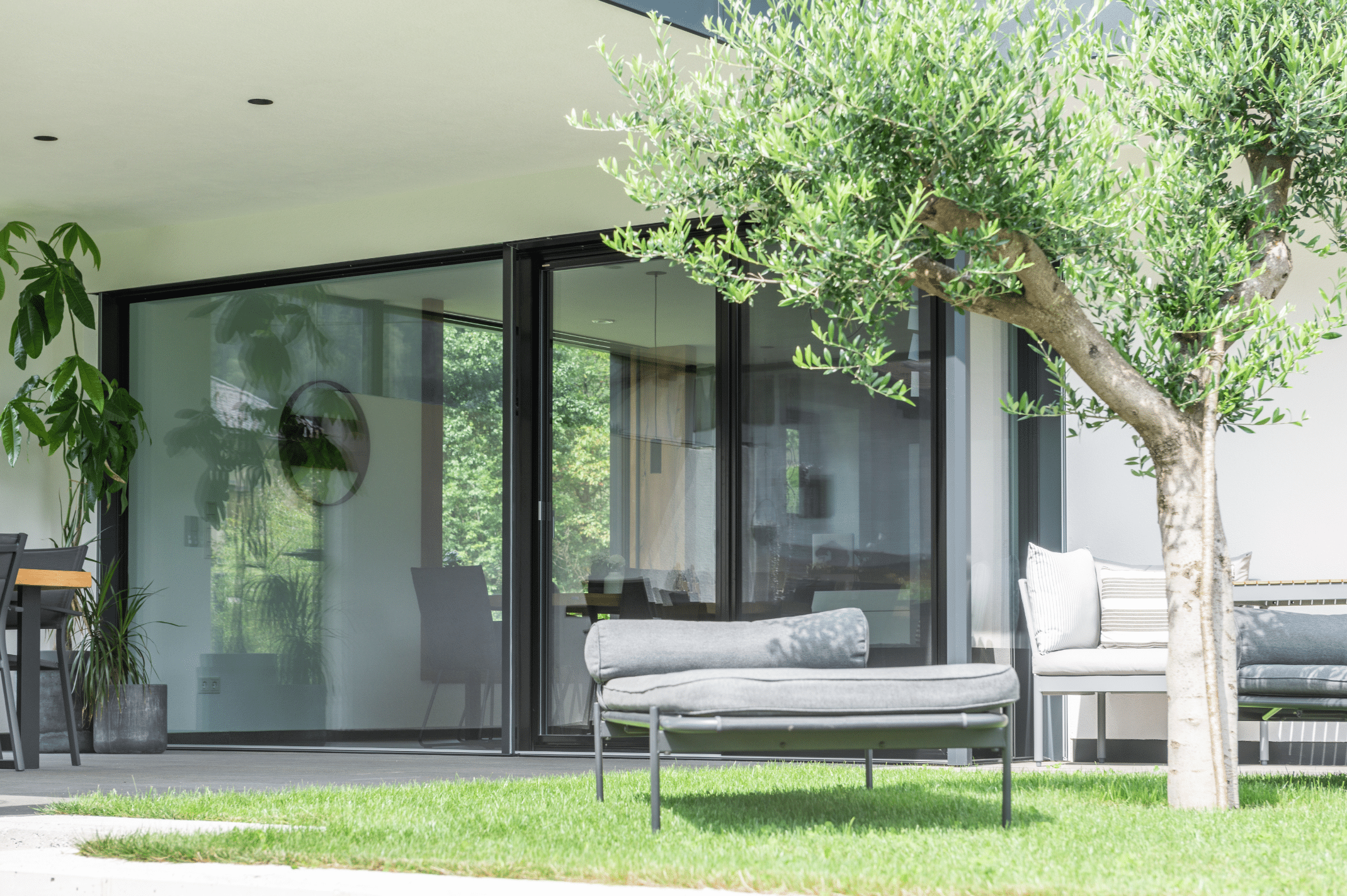 Internorm zeigt einen Garten mit gemütlichem Sonnenbett, einer überdachten Terrasse und großen Glasfronten und einer Schiebetür mit Zugang zum Wohnzimmer.
