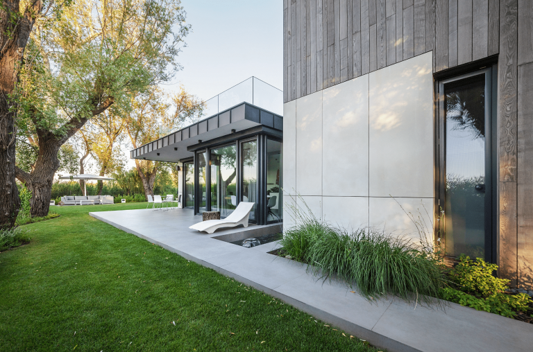 Internorm zeigt die Rückseite eines modernen Holzhauses mit einer Dachterrasse und einem Garten mit gefliester Terrasse und großen Glasfronten beim Zugang zu dem Wohnbereich.