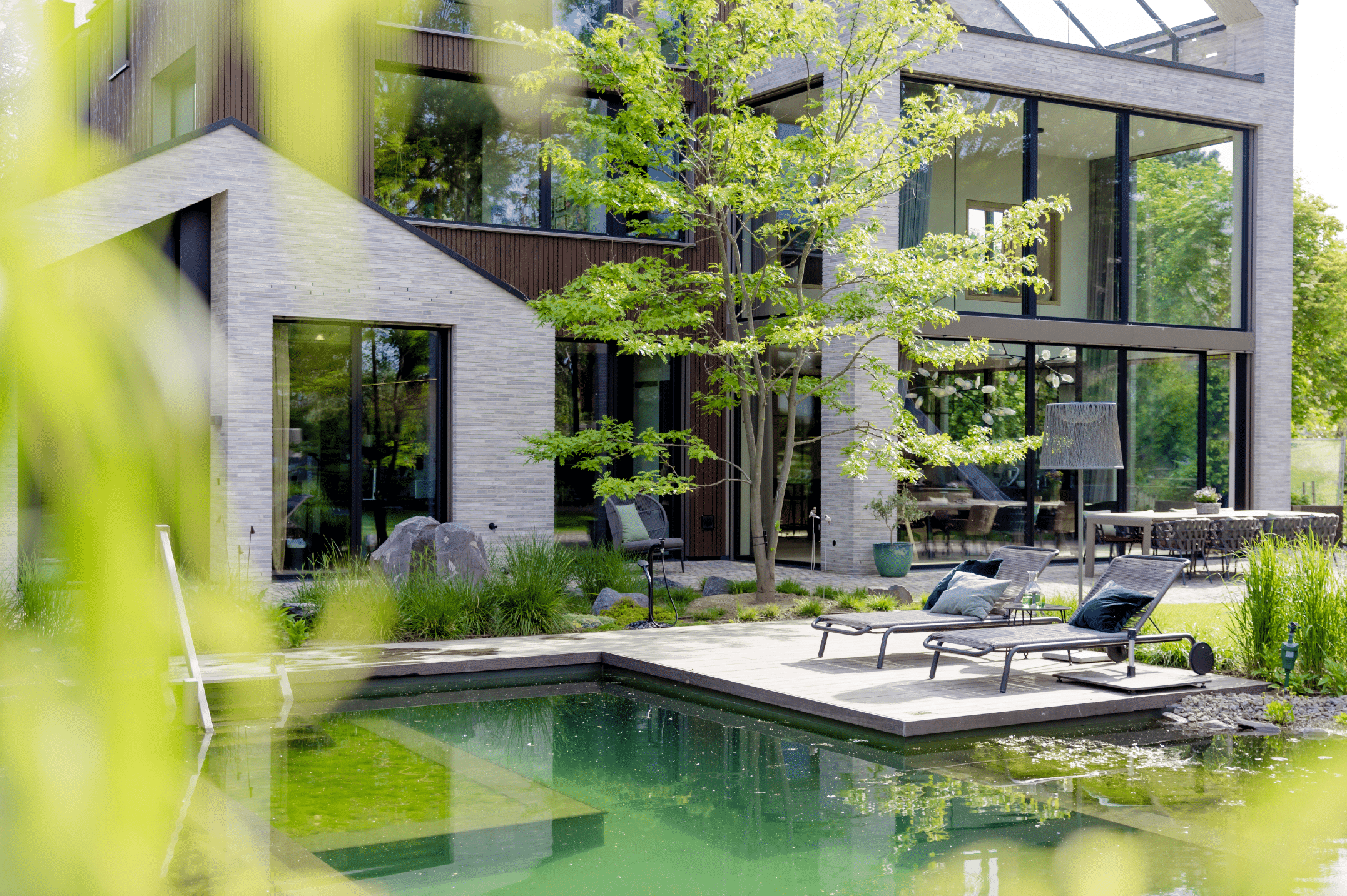 Josko zeigt ein modernes Haus mit vielen Fenstern und großen Glasfronten und einem Garten mit natürlichem Pool, vielen Pflanzen und einem Sonnendeck mit Liegen daneben.