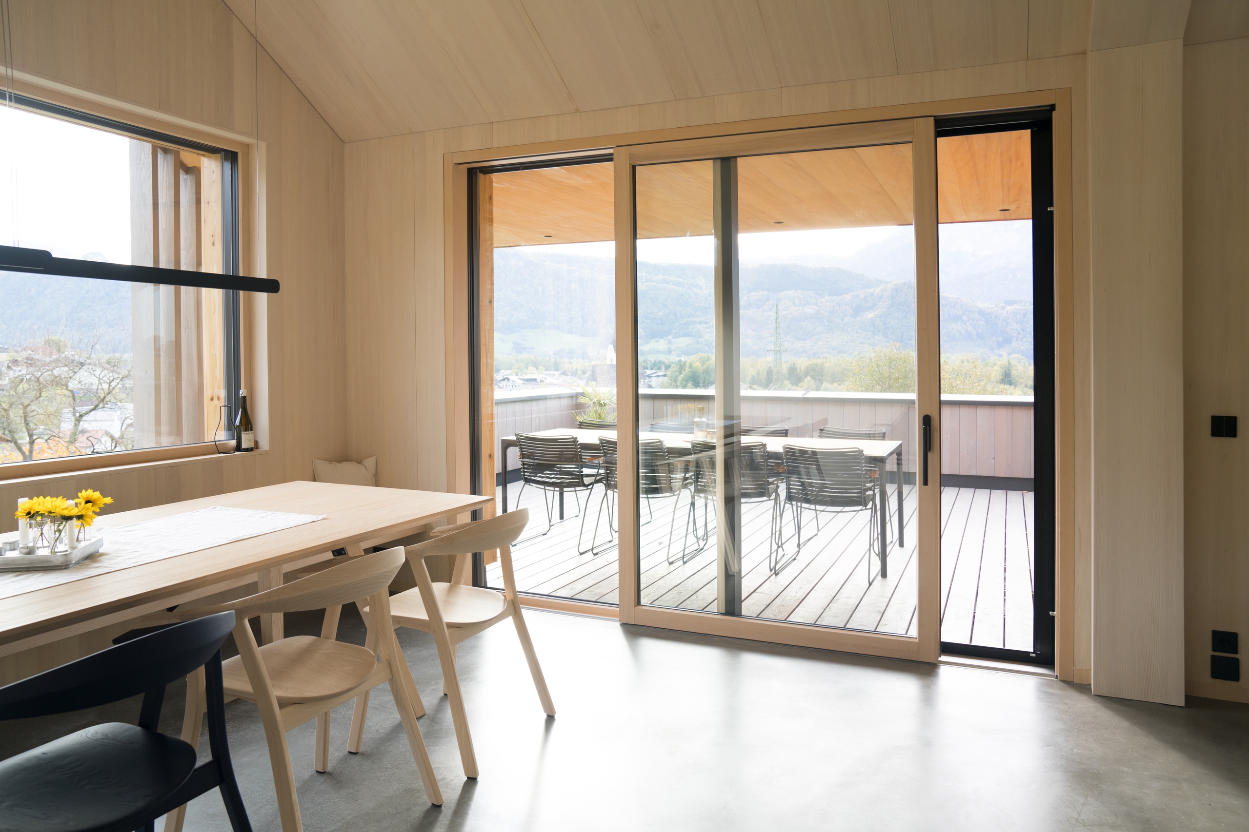 Heller Wohnraum mit Glasfront und Glasschiebetüre mit hellbraunem Rahmen von Josko in einem Holzhaus mit Blick auf die Terrasse.