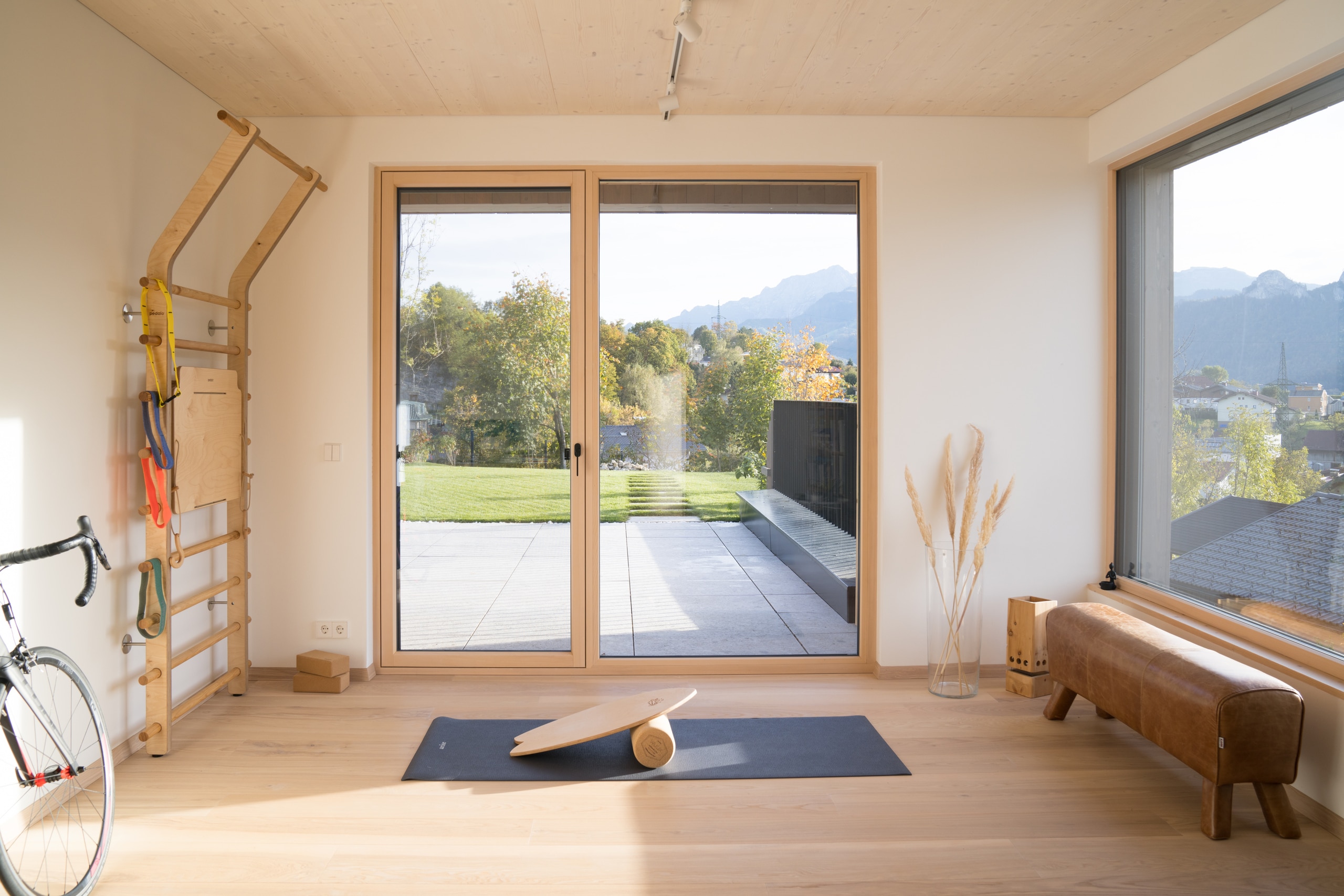 Heller Fitnessraum mit großem fix verglasten Fenster, Glasfront und Glastüre mit hellbraunem Rahmen von Josko mit Blick auf Terrasse und Garten.