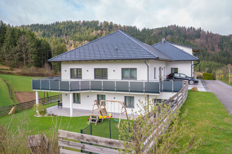 Doppelhaus mit anthrazitfarbenem Satteldach, Balkongeländer aus dunklem Glas und überdachtem Terrassenbereich.