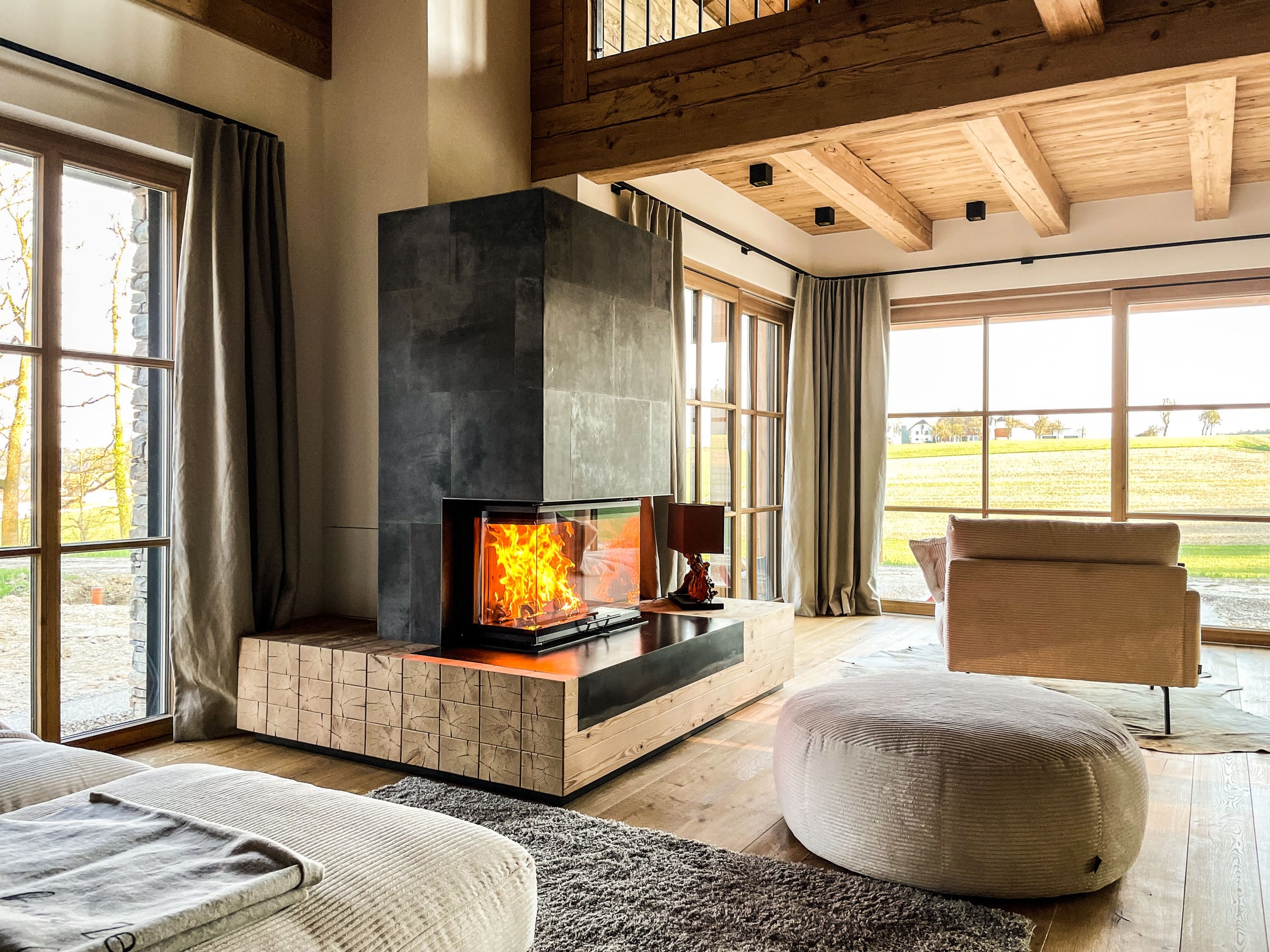 Offener Wohnraum mit Holzdecke und Kamin mit zurückversetztem Sichtfenster, grauen Steinplatten auf Sockel aus quadratischen Holzbalken von Ofenmanufaktur Liebewein.