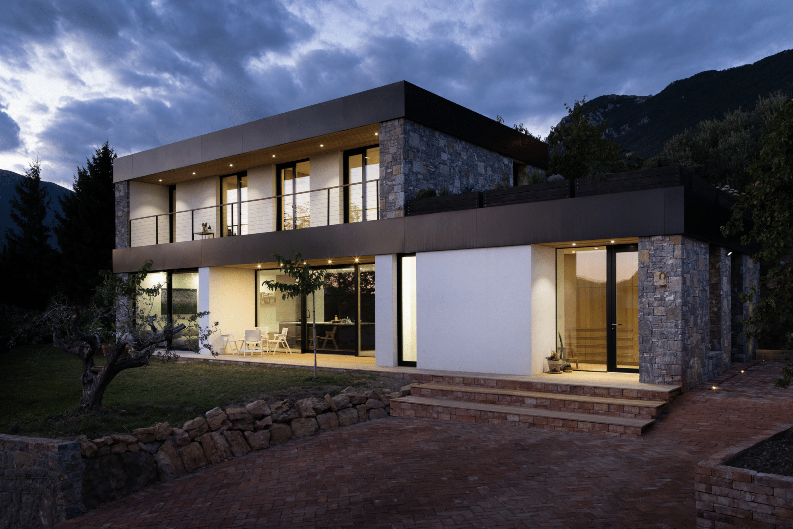 Modernes Flachdach-Wohnhaus bei Nacht mit integrierter Terrasse, bodentiefer Fixverglasung, Hebeschiebetür und Glas-Eingangstüre mit schwarzem Rahmen und rahmenlosen Glasseitenteil von M SORA.