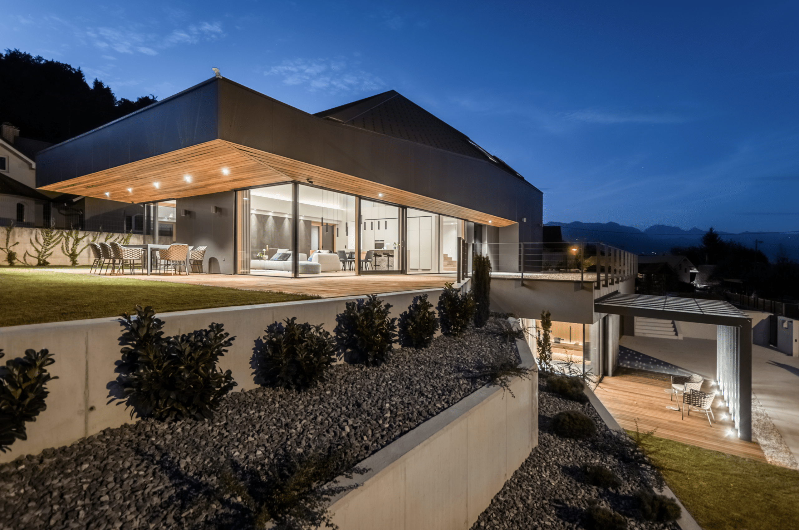 Modernes Wohnhaus bei Nacht mit überdachter Holzterrasse, bodentiefer Fixverglasung und Hebeschiebetür mit schwarzem Rahmen von M SORA.