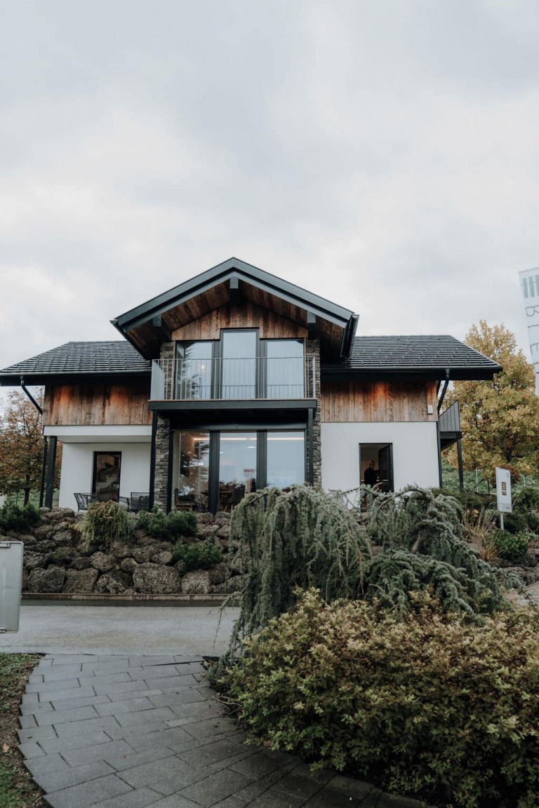 Musterhaus mit Zwerchdach, Balkon, überdachter Terrasse und einem Fassadenmix aus Beton, Holz und Stein.