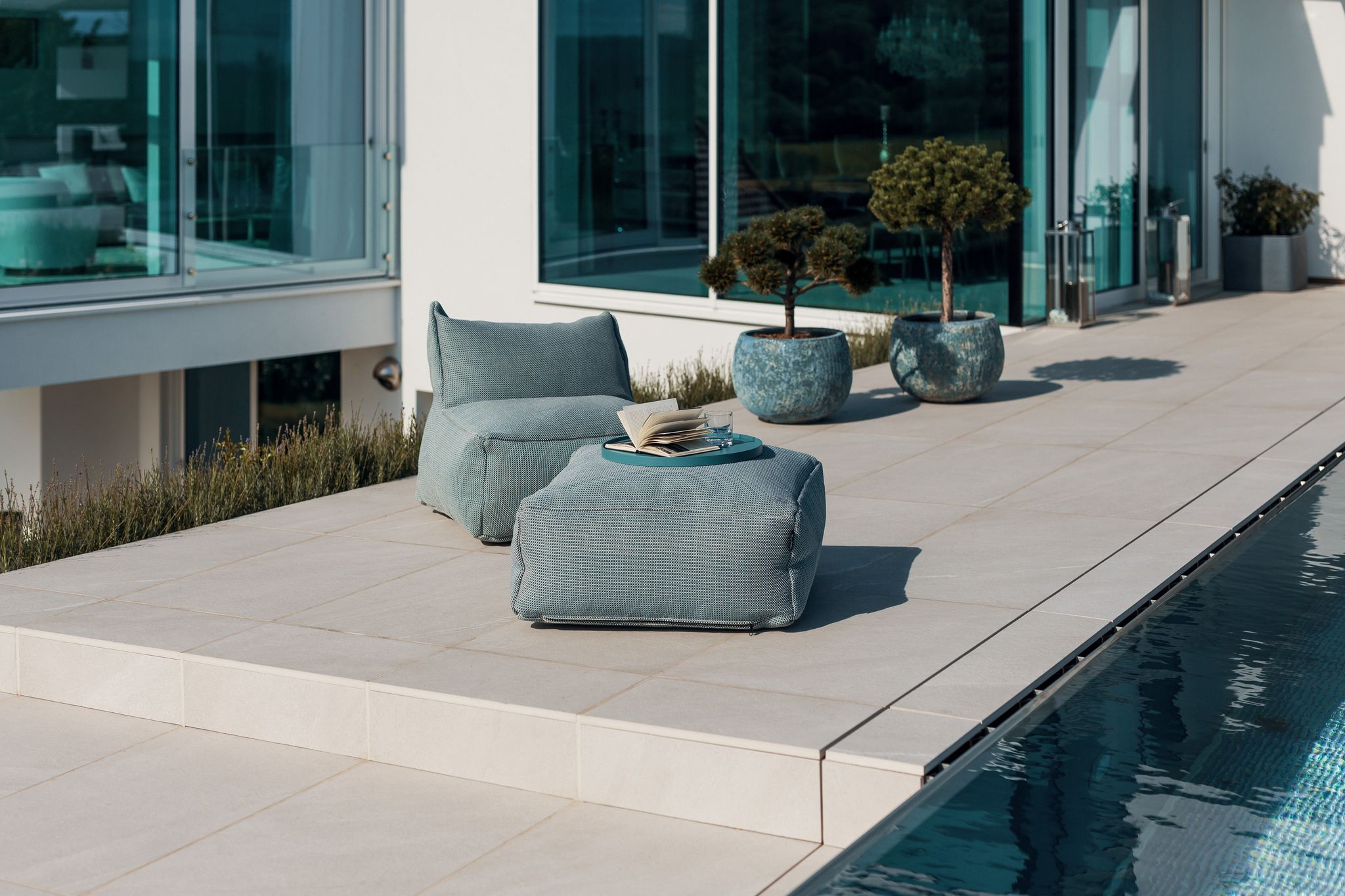 Nentwich zeigt auf diesem Foto eine geflieste Terrasse, einen Pool, Sitzpouf mit Tisch und ein schönes Haus mit vielen Glaselementen im Hintergrund.