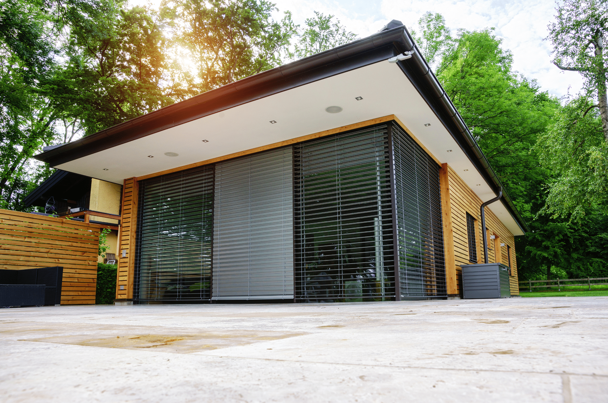 Newo Sonnen- und Insektenschutz zeigt ein Poolhaus mit Flachdach und Holzverkleidung an dessen Terrassentüren schwarze Rollläden und ein grauer Raffstore montiert sind um vor der Sonne zu schützen.