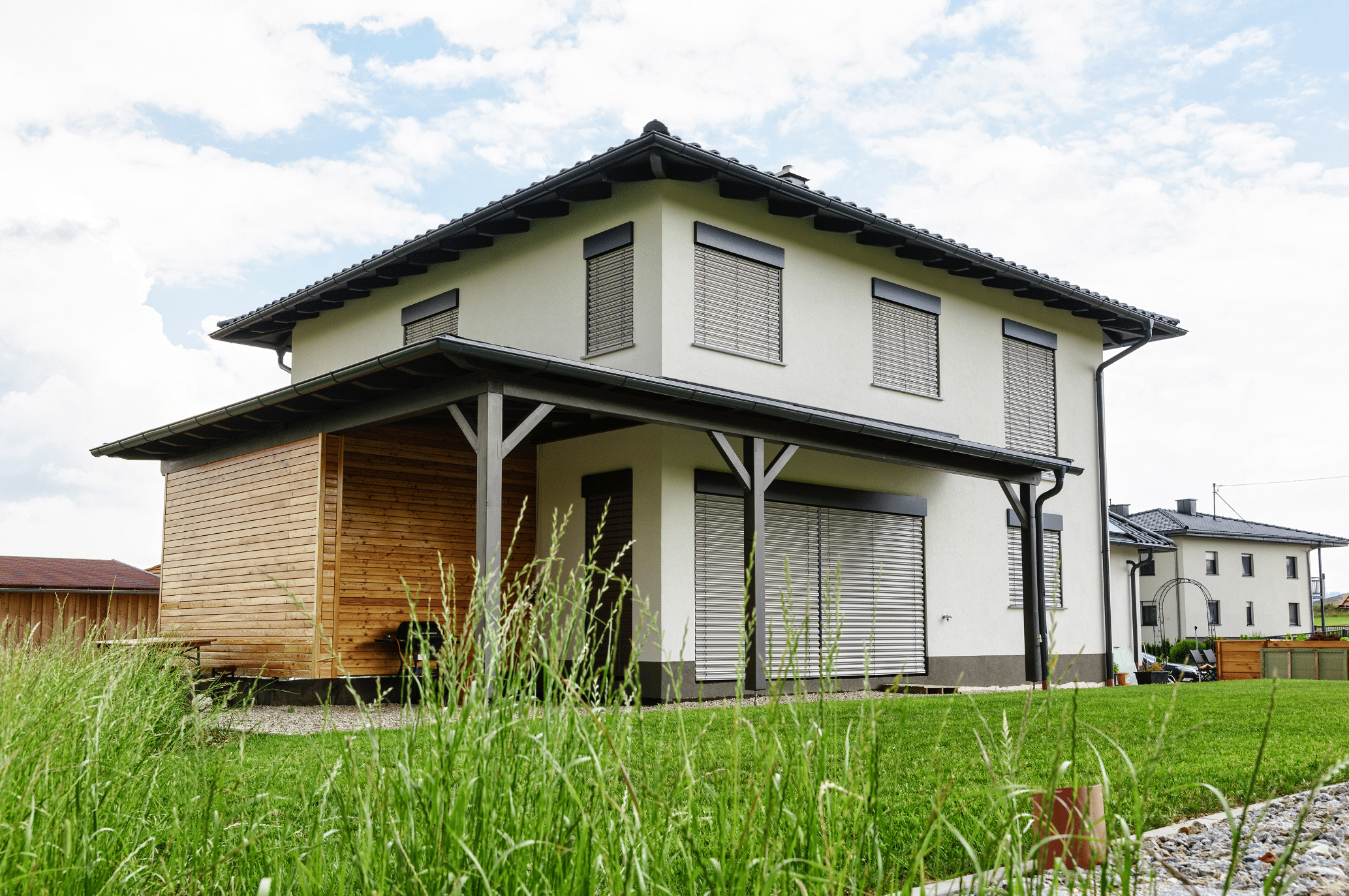 Newo Sonnen- und Insektenschutz zeigt ein Einfamilienhaus mit vielen Fenstern an denen ein grauer Raffstore angebracht ist und einen Anbau mit Holzverkleidung neben der überdachten Terrasse.