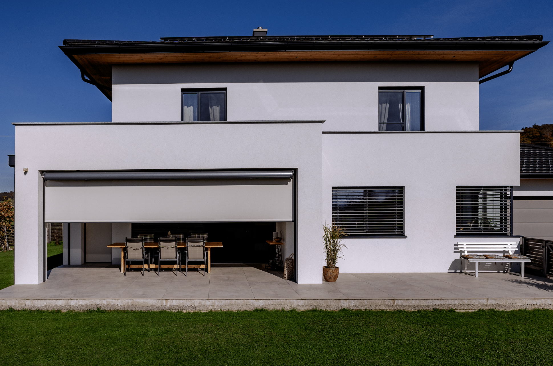 Newo Sonnen- und Insektenschutz zeigt die überdachte Terrasse eines Einfamilienhauses, mit grauem Textilscreen, Fliesenboden und einer gemütlichen Sitzgruppe bestehend aus einem Holztisch und Stühlen.