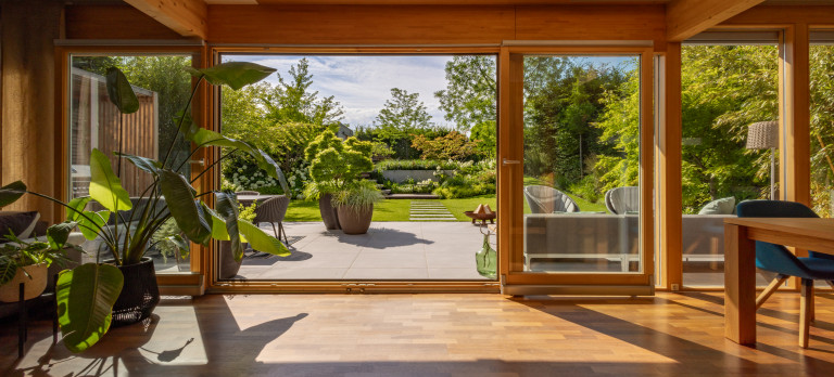 Praskac Pflanzenland Prachtgarten zeigt lichtdurchfluteten Wohnraum mit großer Zimmerpflanze mit Blick auf die Terrasse und Gartenanlage mit Trittsteinen, Stufen und Hochbeet.